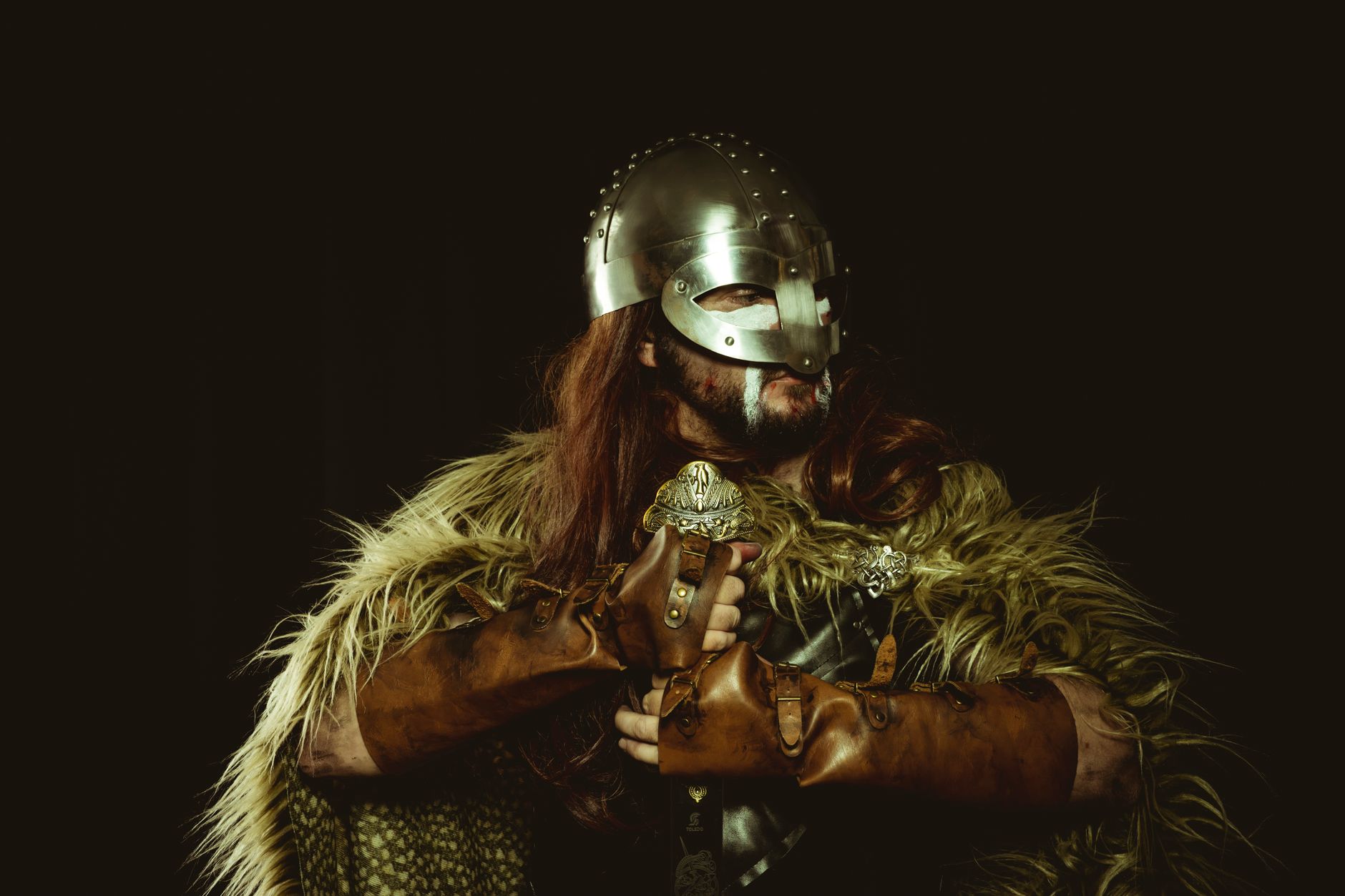 A viking warrior wearing a helmet and armor with fur covering against a dark background.