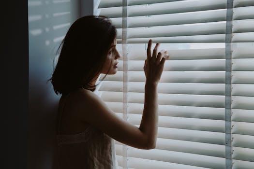A young woman in a nightdress gazes thoughtfully through window blinds, creating a serene and introspective mood.