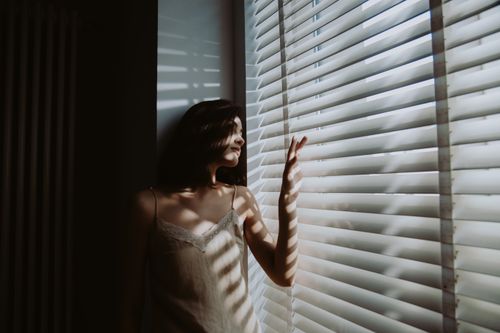 Free A woman stands in a dimly lit bedroom, gazing through window blinds as morning light casts shadows. Stock Photo