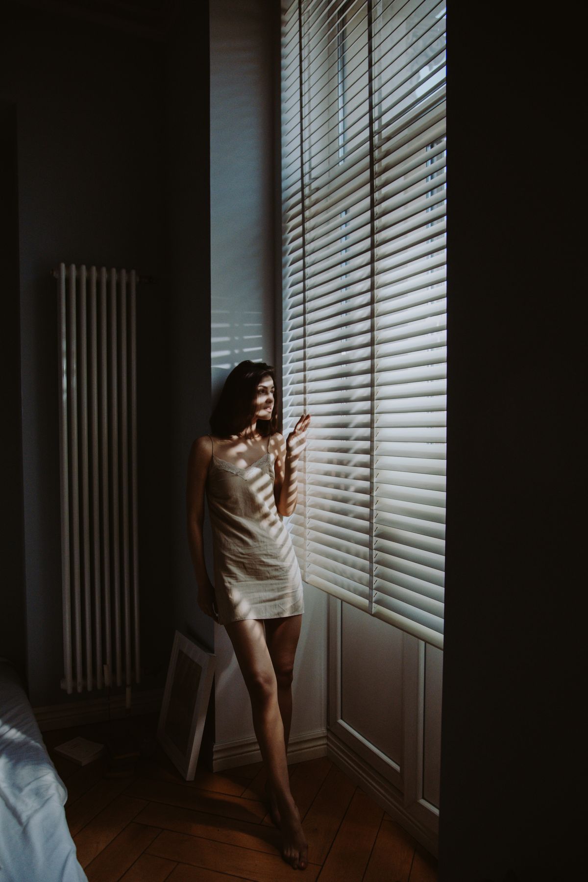 A woman stands by a window, embraced by gentle morning light filtering through shutters, in a cozy bedroom.