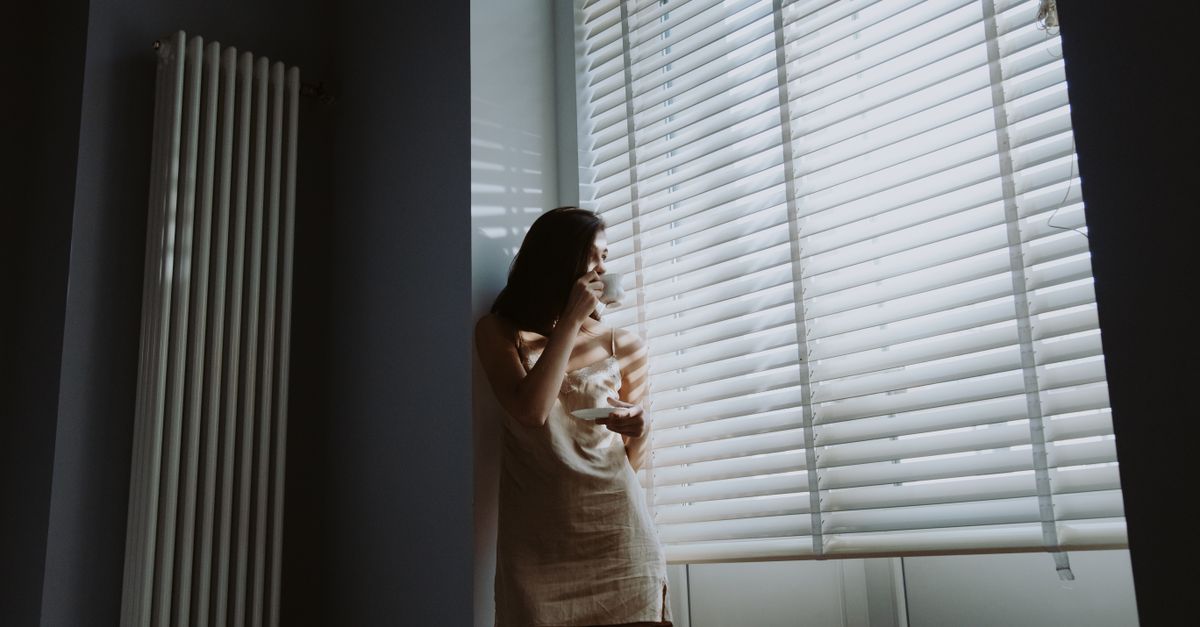 A woman in soft morning light stands by the window, holding a cup and saucer, savoring a peaceful moment.