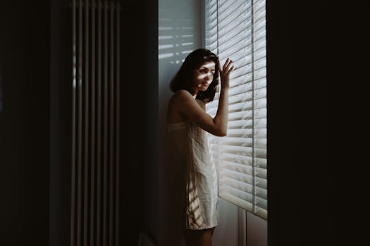 A woman in sleepwear peeks through window blinds, bathed in soft, morning light.