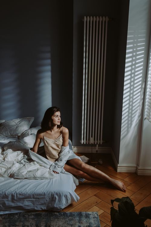 Free Woman sitting in bed, gazing out a window with morning light casting shadows in a cozy bedroom. Stock Photo