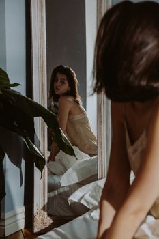 A woman in nightdress admires her reflection in a bedroom mirror, surrounded by soft natural light.
