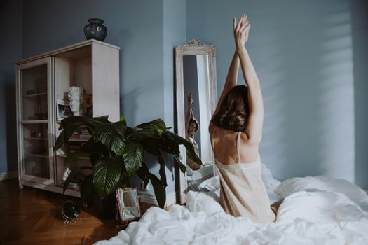 A woman in sleepwear stretches in a calm bedroom setting, reflecting in a mirror.