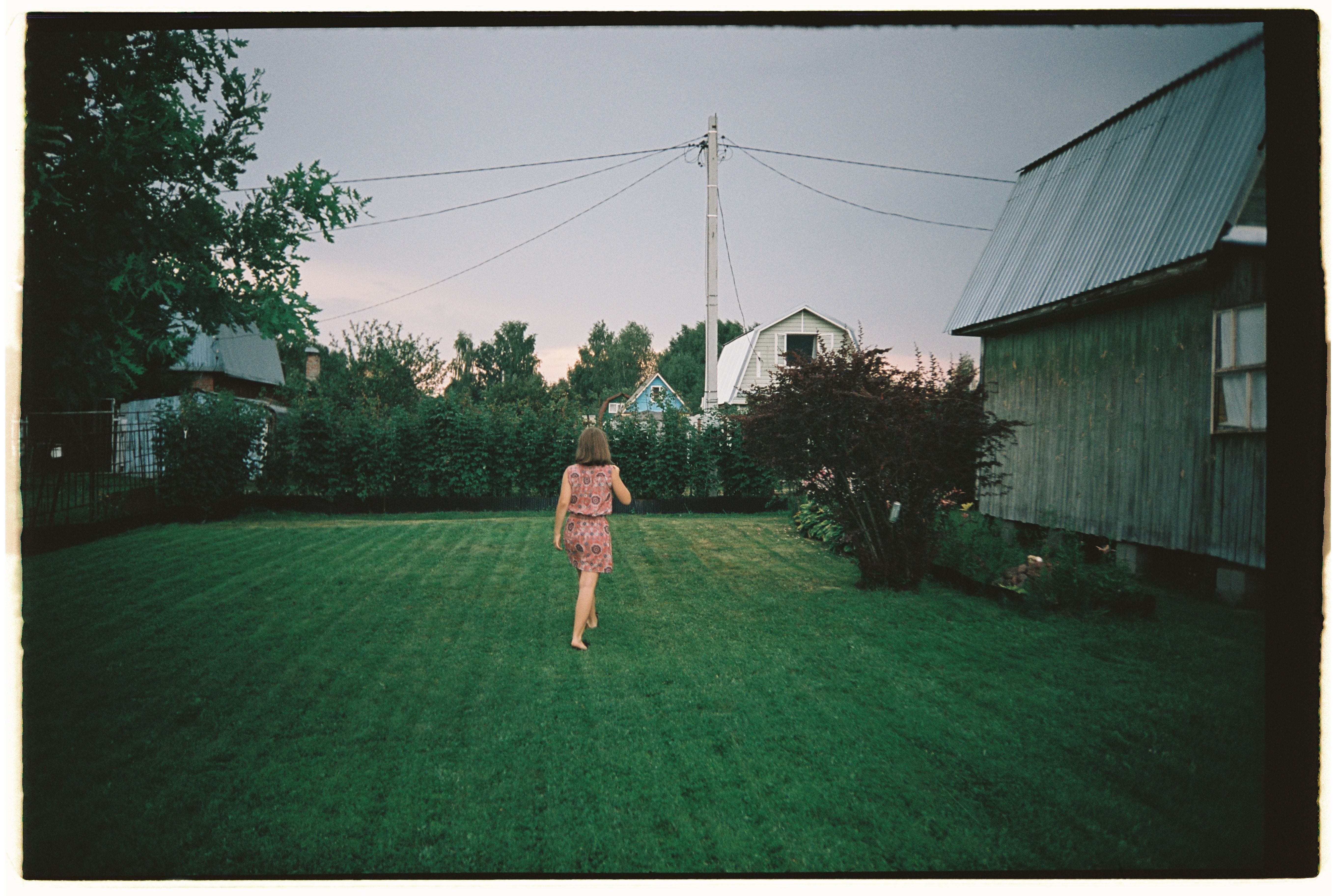A girl walks across a rural summer garden with houses in the background.