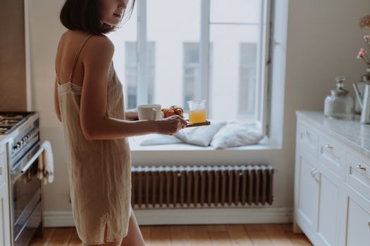 A woman in nightwear holds a breakfast tray with coffee and juice in a bright kitchen.