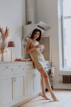 A young adult woman enjoys a morning coffee in a cozy, sunlit kitchen with modern decor.