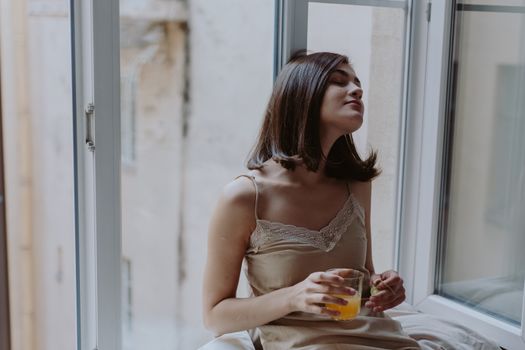 Woman in a spaghetti strap dress savoring orange juice on a window sill, indoors.