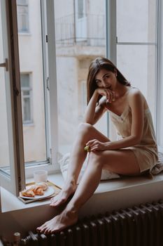 A woman in a casual outfit enjoying a croissant while seated on a windowsill in a cozy indoor setting.