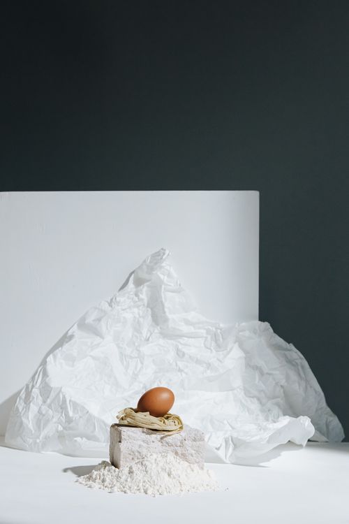 Free A minimalist composition featuring flour, an egg, and pasta against a textured backdrop. Stock Photo
