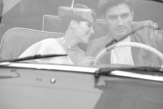 Elegant couple in vintage car, captured in timeless black and white, exuding romance and style.