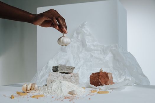 Artistic composition featuring a hand arranging garlic with pasta ingredients in a studio setting.