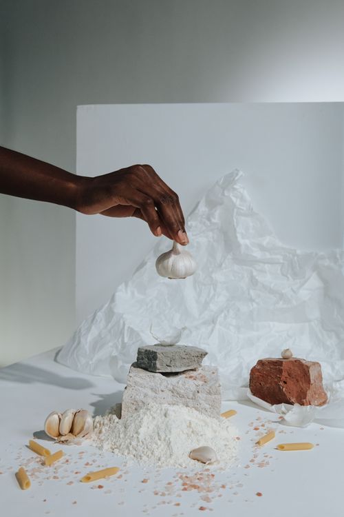 Free Creative still life featuring a hand holding garlic over flour with pasta and rocks. Stock Photo