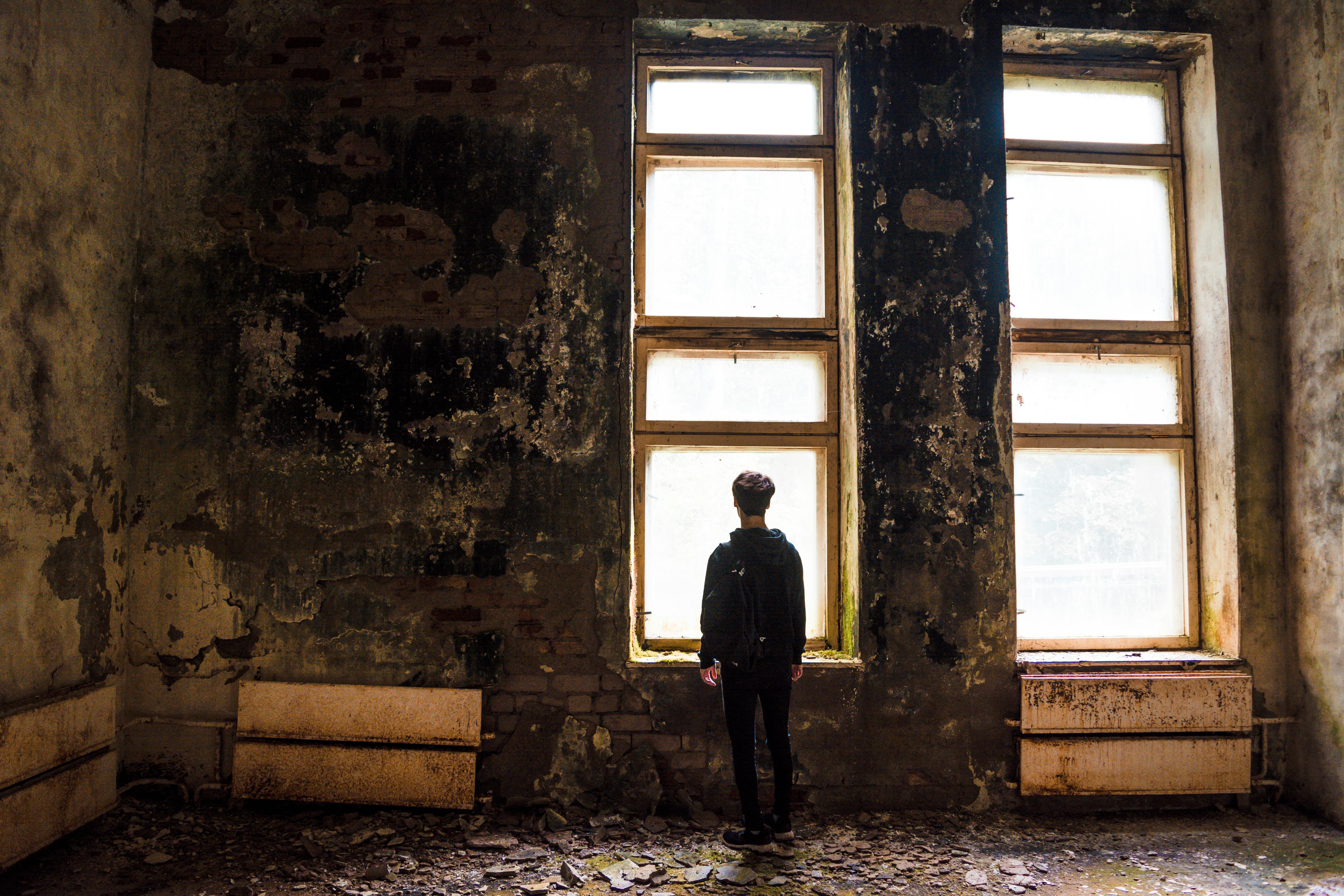 A solitary figure stands facing windows in a dilapidated urban setting, evoking themes of abandonment.