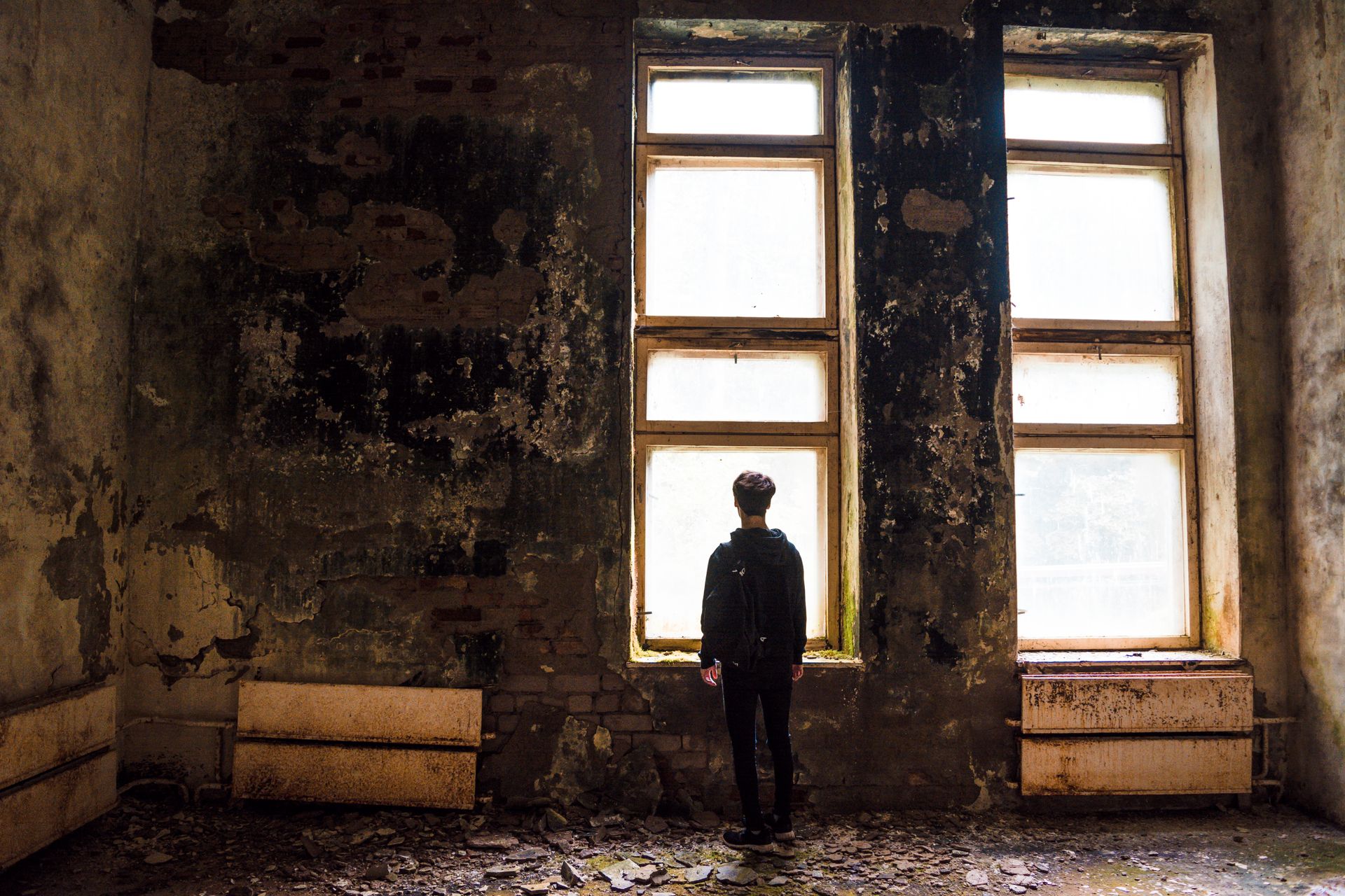 A solitary figure stands facing windows in a dilapidated urban setting, evoking themes of abandonment.