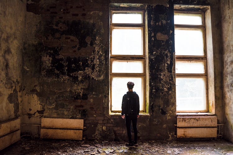 A Back View Of A Man Inside The Abandoned Building While Facing The Window