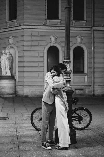 A couple embraces romantically under a lamppost at night in a classical urban setting.