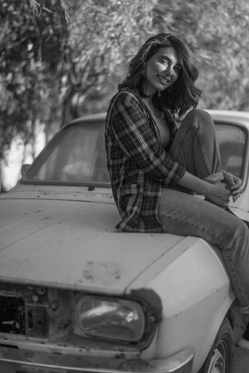A Grayscale Photo of a Woman in Plaid Shirt Sitting on a Car Hood