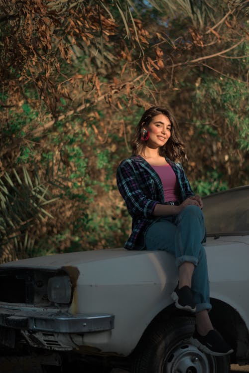 A Woman in Plaid Long Sleeves Smiling while Sitting on a Car Hood