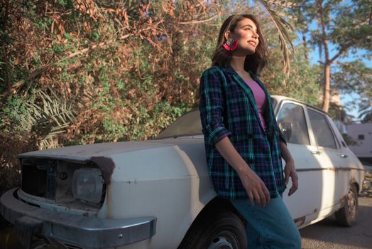 A stylish woman with statement earrings poses confidently against a vintage car in a sunlit outdoor setting.