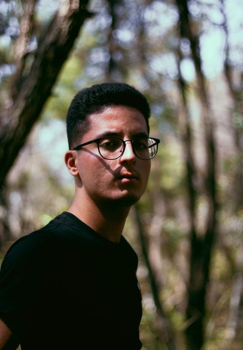 Free Portrait of a young man with glasses in a forest setting under natural sunlight. Stock Photo