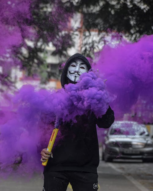 A Person in a Hacker Mask Holding a Smoke Bomb with Purple Colored Smoke