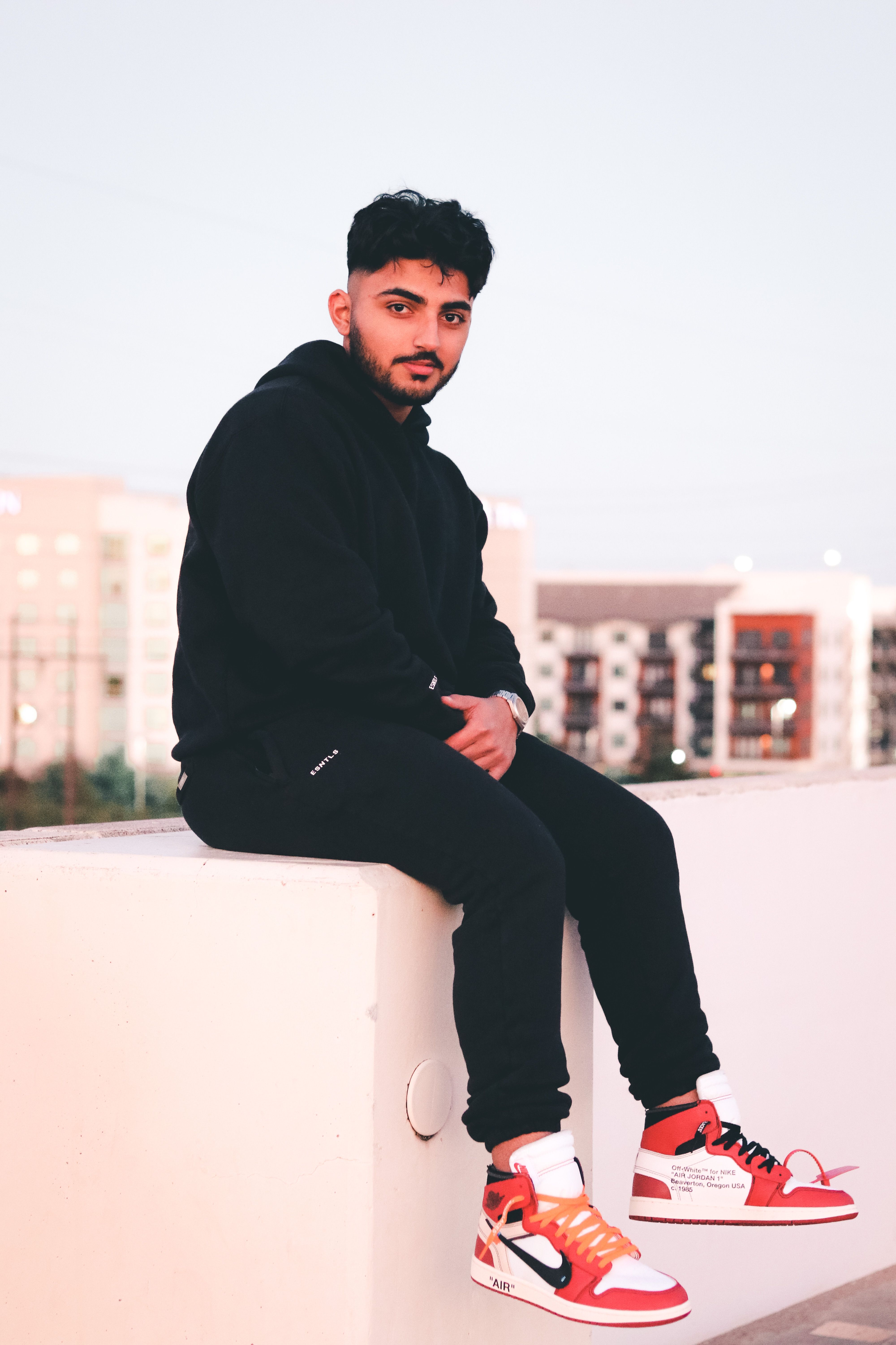 Fashionable man in black outfit sitting on ledge, showcasing streetwear style.