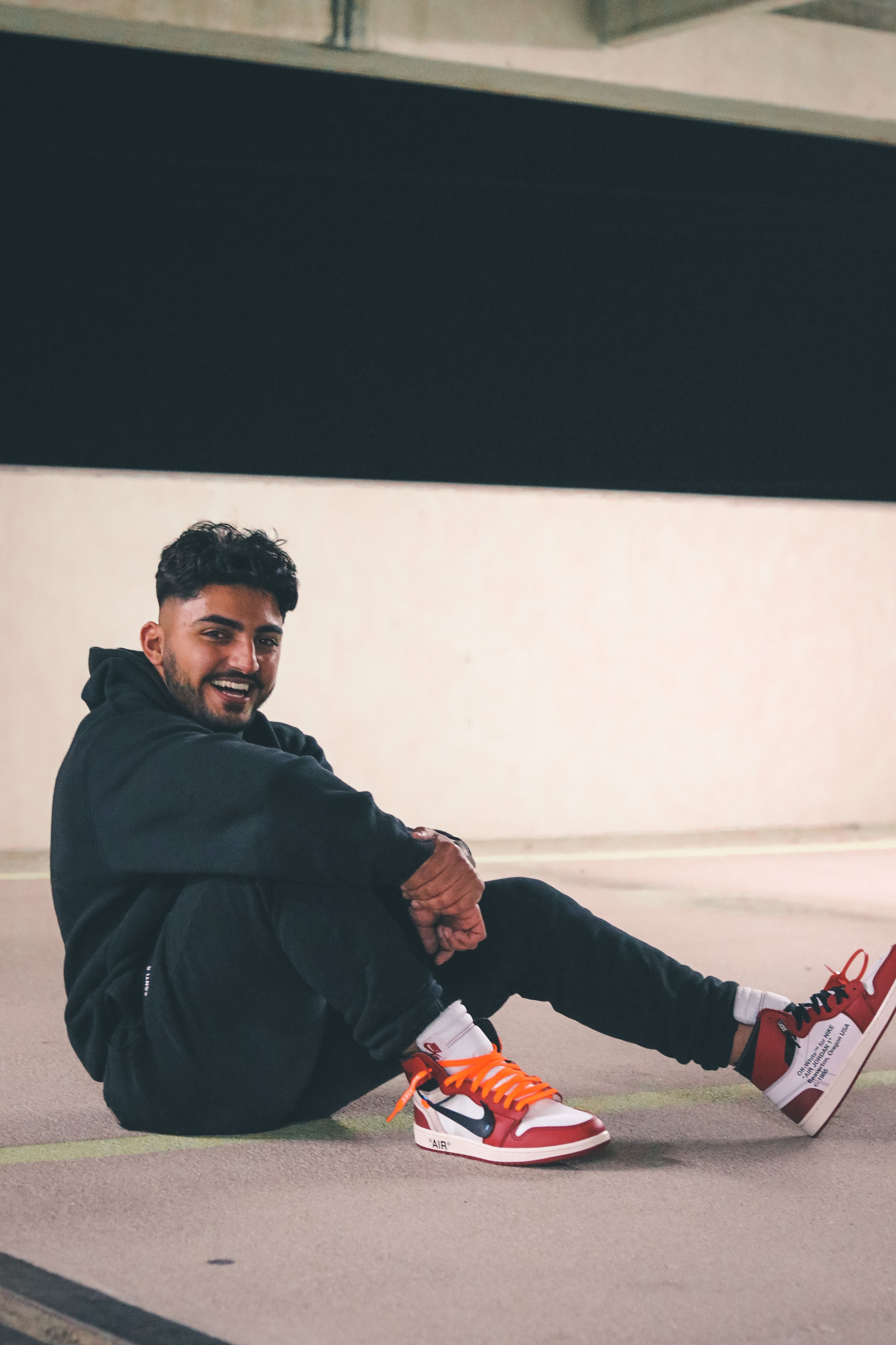 Candid shot of a young man sitting in a black outfit with a smile in urban setting.