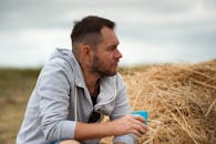 一位留着胡子的男子悠闲地坐在乡村田野的干草堆旁，喝着饮料