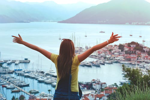 Free Woman Raising Her Hands Facing Cityscape Near Body of Water Stock Photo