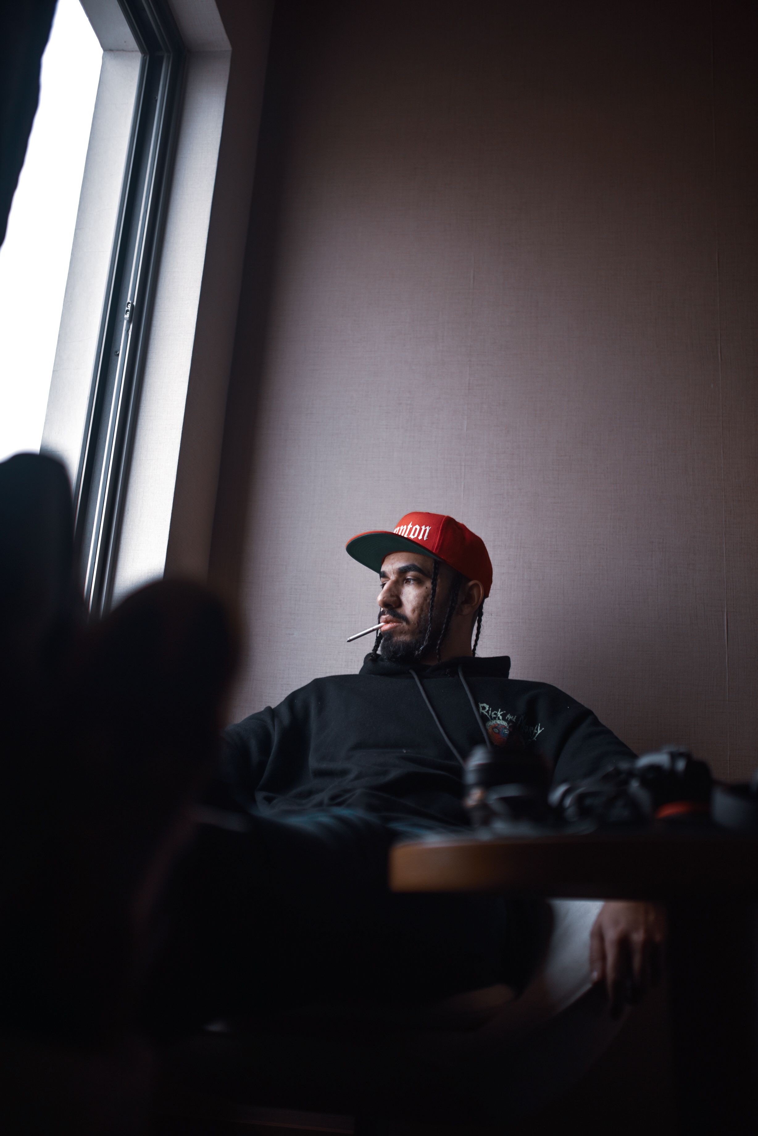 Man wearing a red cap and black sweater sits indoors by a window, smoking.