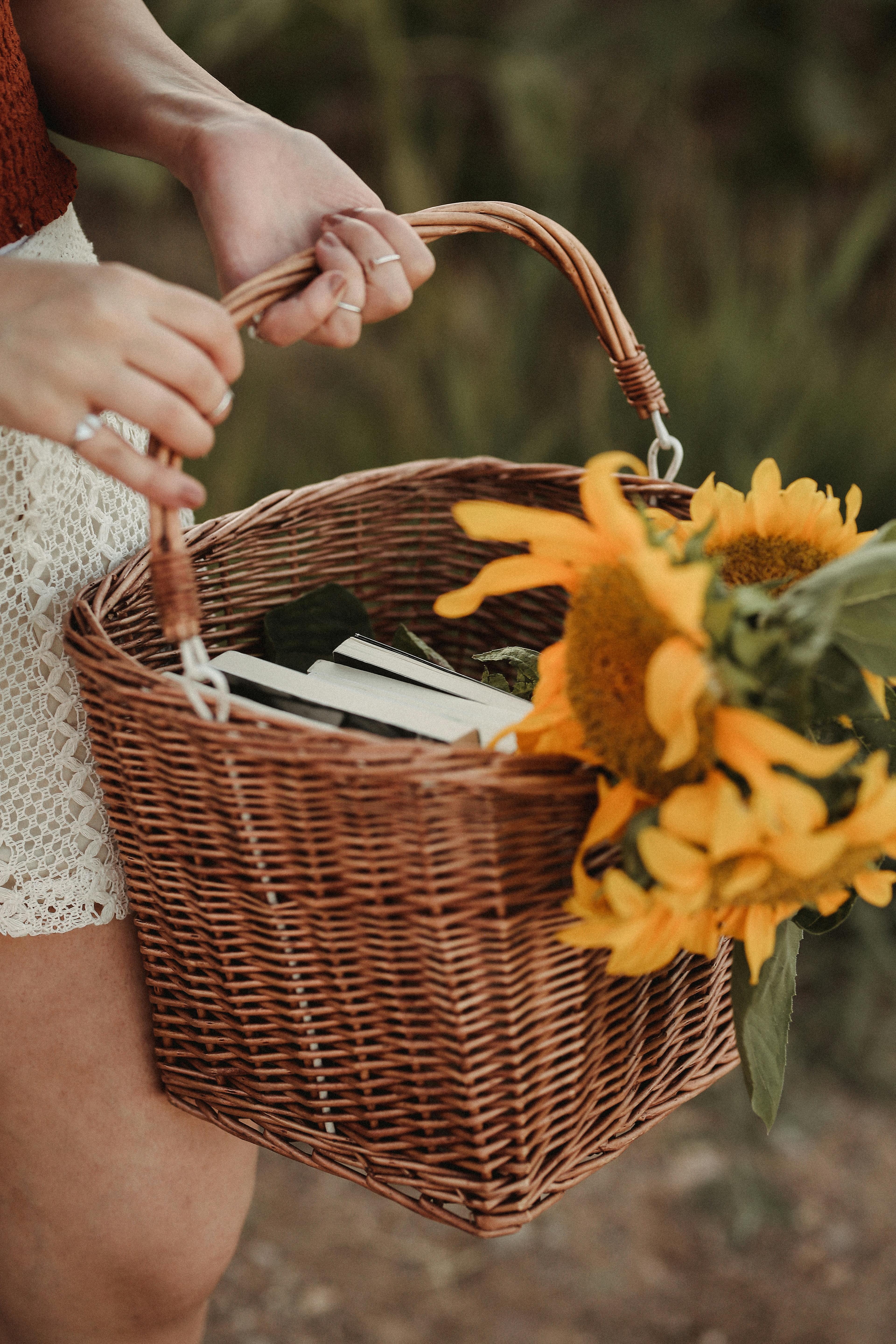 Close-up of a woman holding a wicker basket filled with sunflowers and books in a natural setting.