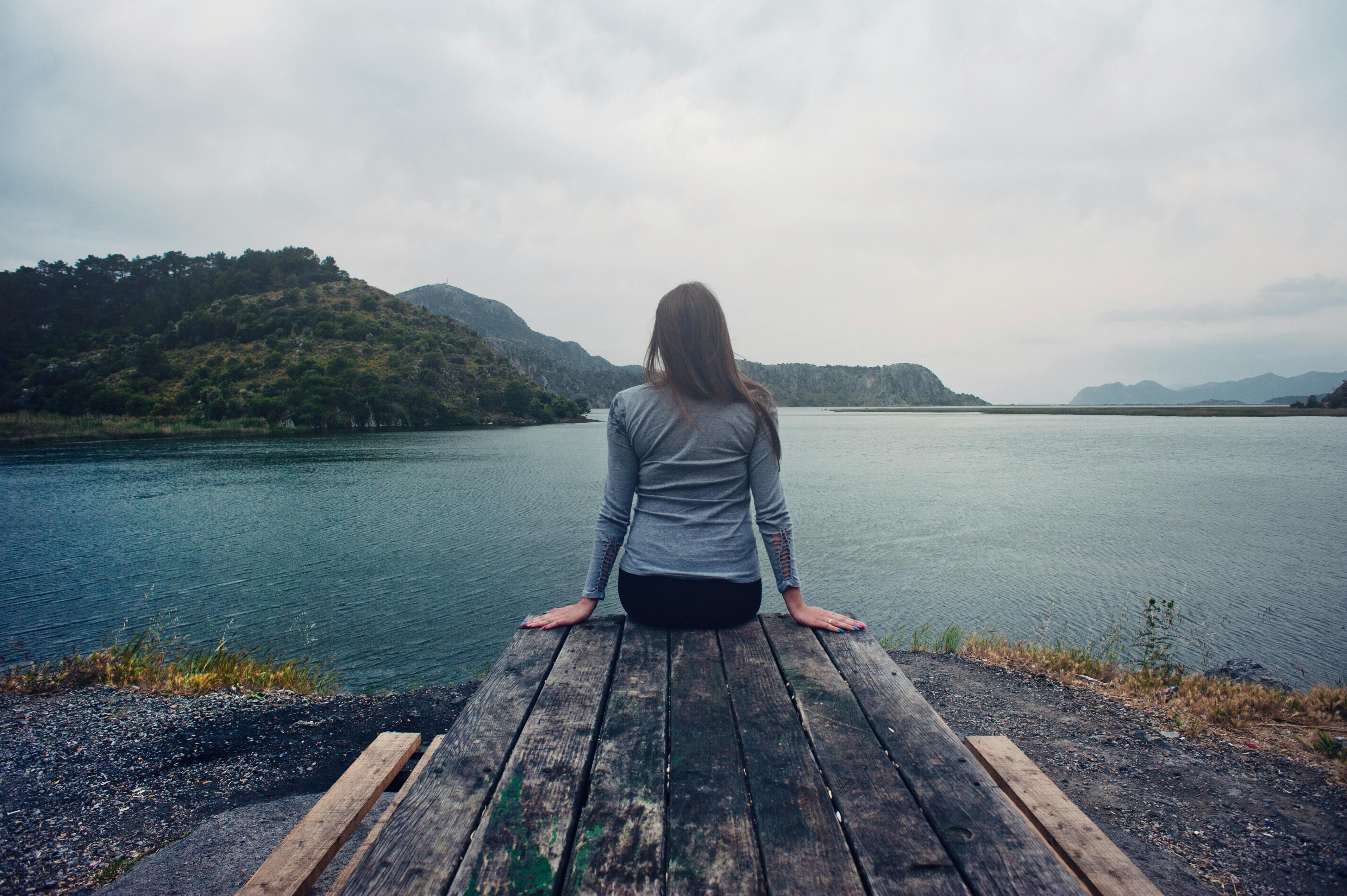 Woman Wearing Gray Long-sleeved Shirt and Black Black Bottoms Outfit  Sitting on Gray Wooden Picnic Table Facing Towards Calm Body of Water at  Daytime · Free Stock Photo