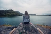 Woman Wearing Gray Long-sleeved Shirt and Black Black Bottoms Outfit Sitting on Gray Wooden Picnic Table Facing Towards Calm Body of Water at Daytime