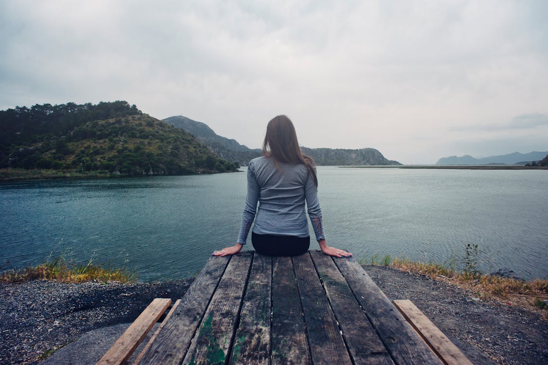 Vrouw Draagt Grijs Shirt Met Lange Mouwen En Zwarte Zwarte Bodems Outfit Zittend Op Grijze Houten Picknicktafel Geconfronteerd Met Kalm Water Overdag