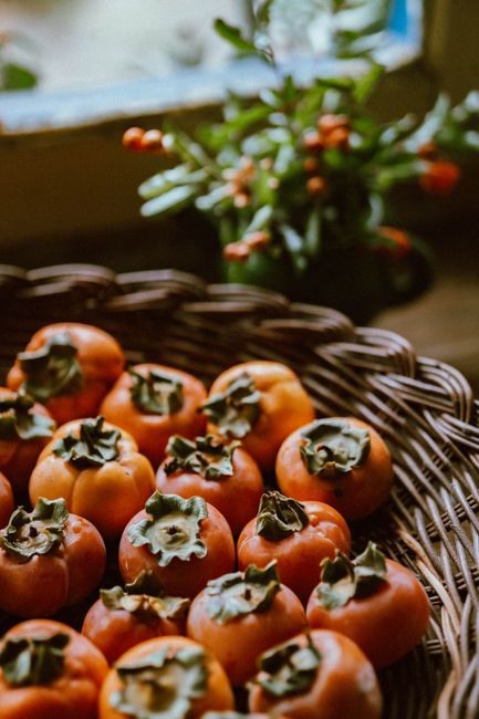 A pile of glossy orange caquis persimmons, one sliced in half showing soft jelly-like orange flesh and dark seeds, next to whole unripe firm ones