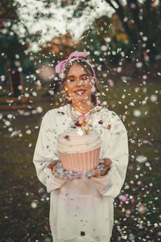 Smiling woman holding a giant cupcake surrounded by confetti in an outdoor setting.