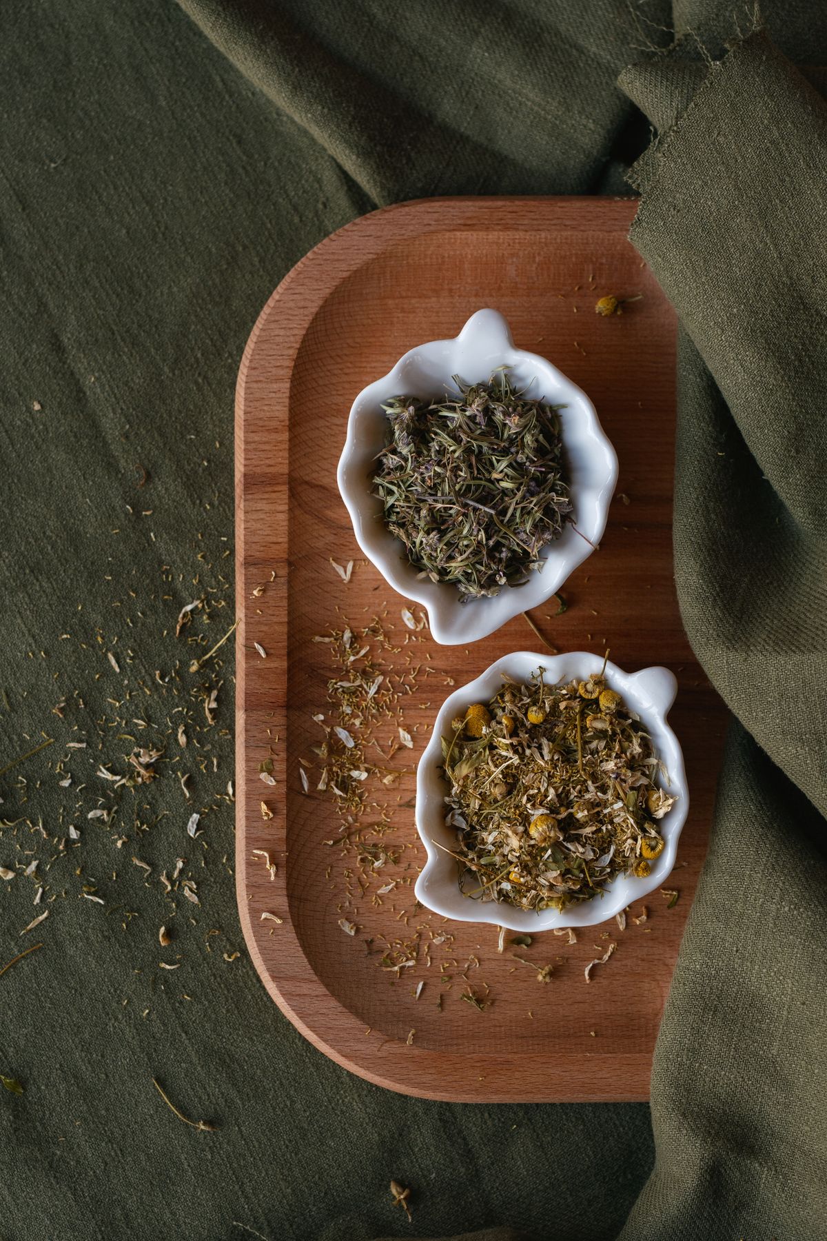 Two small white bowls of dried herbs and chamomile flowers on a wooden tray over a green cloth.