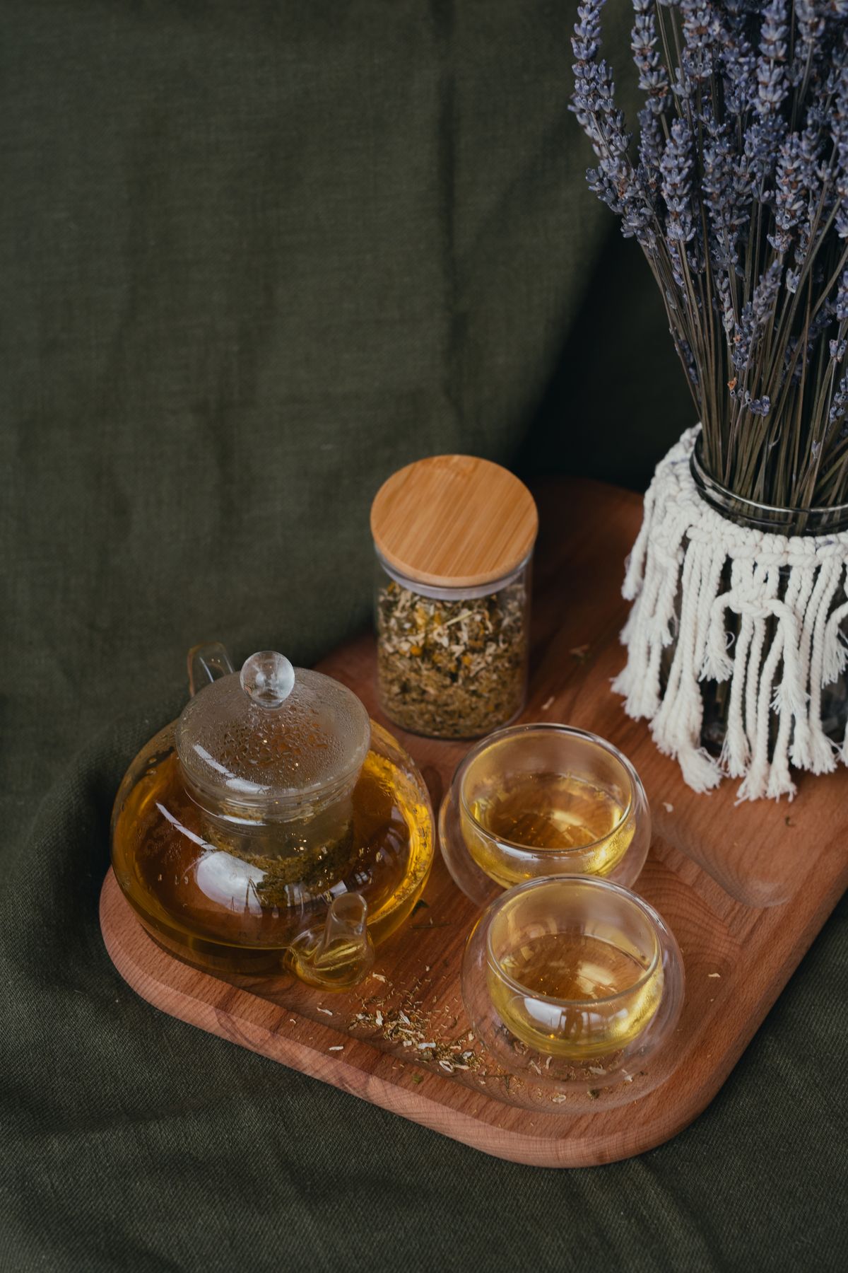 A glass teapot and cups of herbal tea beside dried chamomile and lavender on a wooden tray.