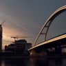 A modern arch bridge spans a wide river at dusk, with construction cranes operating on a high-rise development in the background.