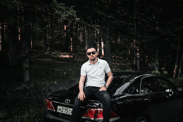 Man In White Polo Shirt Wearing Sunglasses While Sitting On Black Car