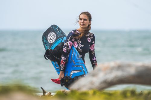 Free Female kitesurfer on a beach holding a blue kiteboard, wearing floral sportswear. Stock Photo