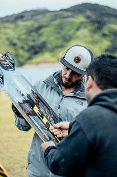 Two men examining a kiteboard outdoors, wearing casual jackets and hats.