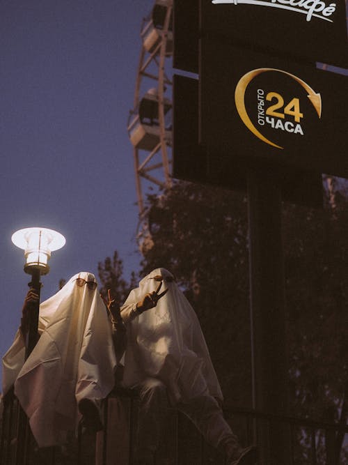 People Covered with White Blankets Sitting Next to Street Lamp 