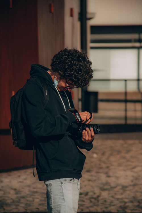Free Young man with curly hair adjusting DSLR camera in a city setting. Natural light photography. Stock Photo
