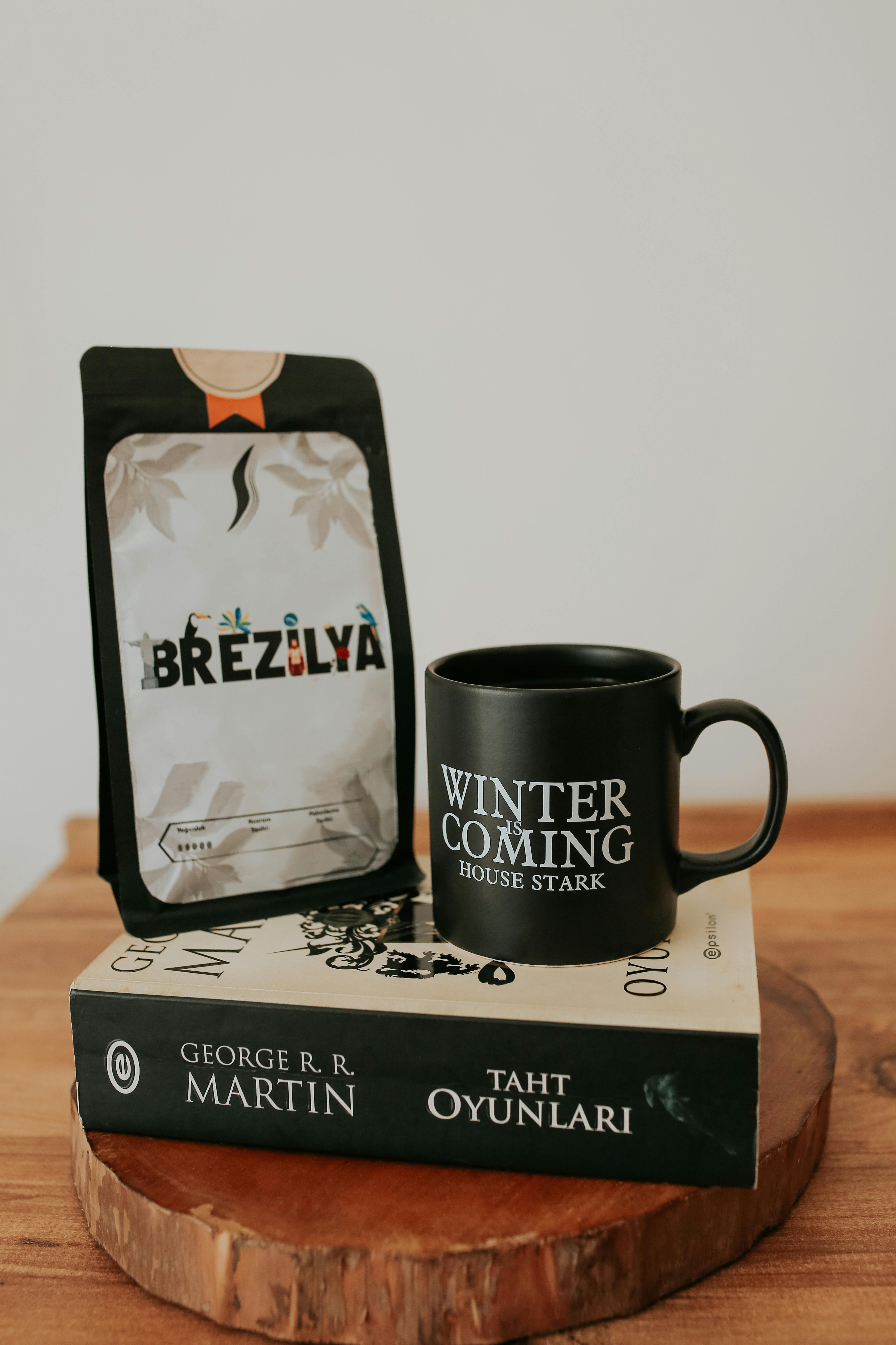 Black mug with 'Winter is Coming' text next to coffee bag on wooden table with book.