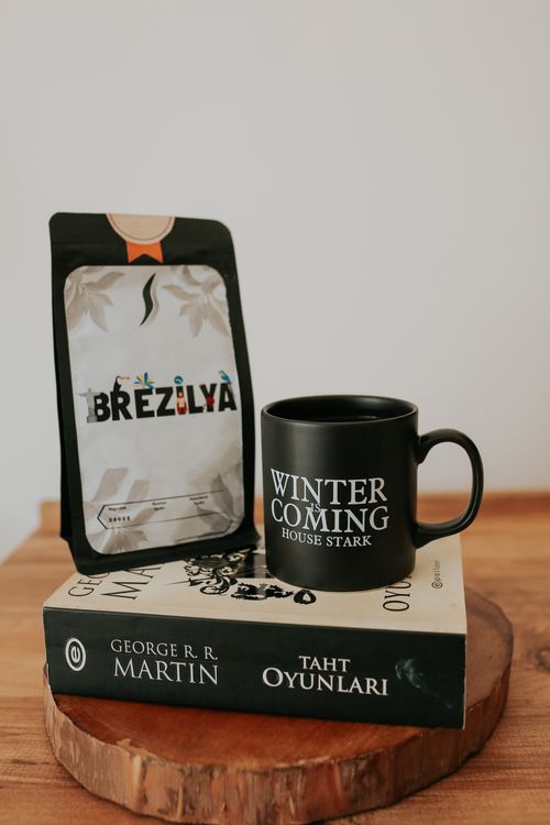 Free Black mug with 'Winter is Coming' text next to coffee bag on wooden table with book. Stock Photo