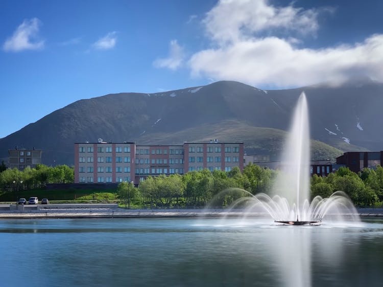 Fountain On Pond, Building And Mountains In Background