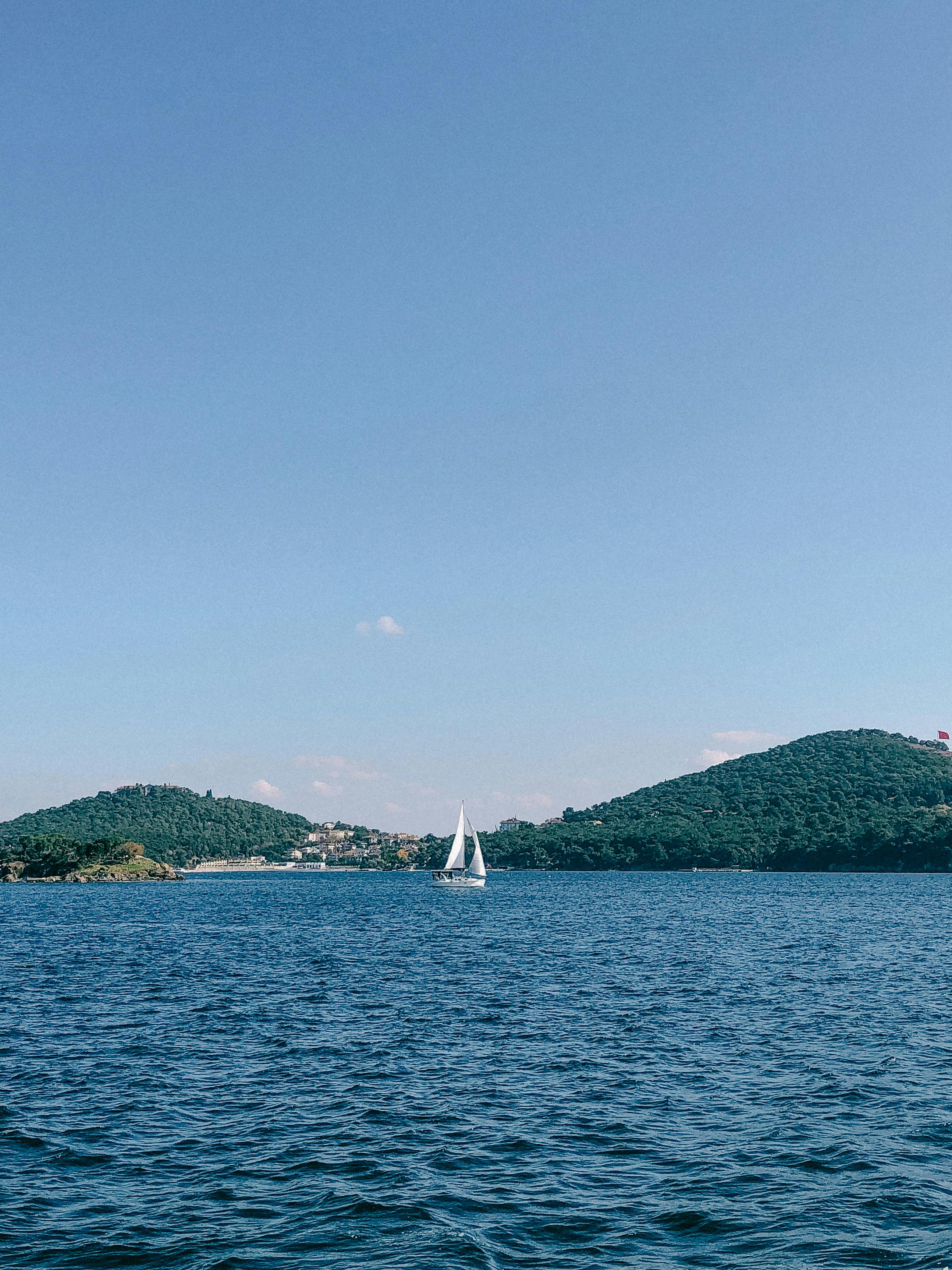 Serene seascape with a lone sailboat against a clear blue sky and green hills.